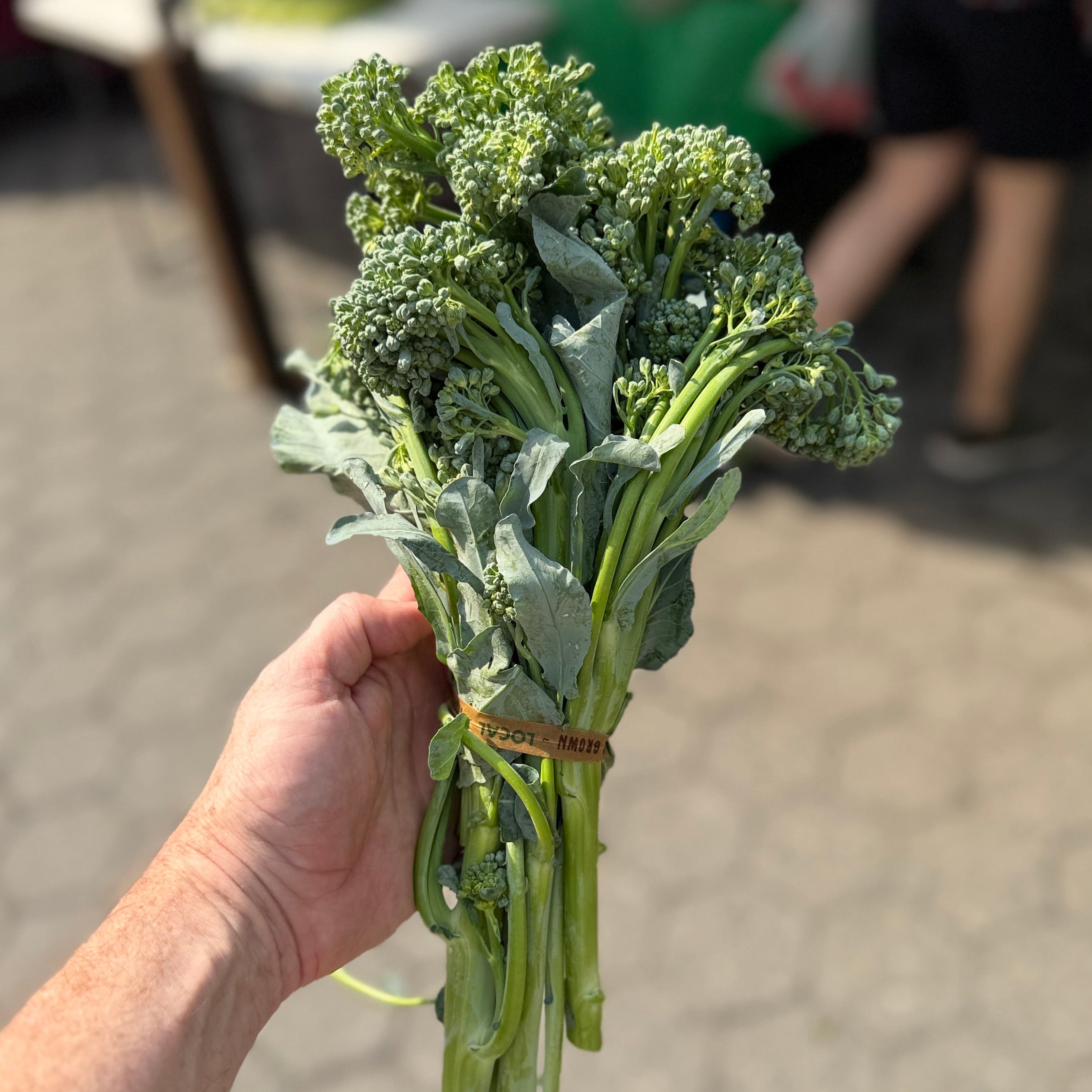 A hand holding a small bunch of fresh green broccoli or brussels sprouts tied with a rubber band, photographed outdoors at what looks like a market with a blurred person in the background.